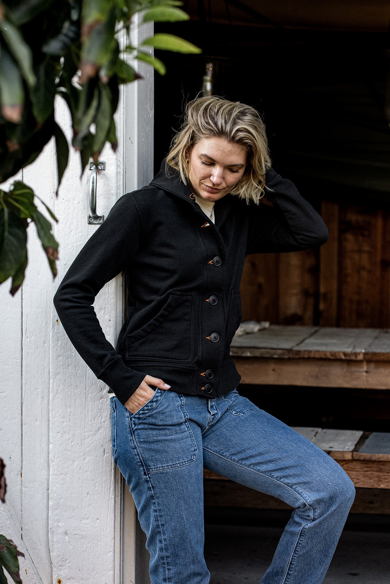 Woman leaning against a white barn door wearing the Bodega shawl French terry cardigan in basalt black with blue jeans.