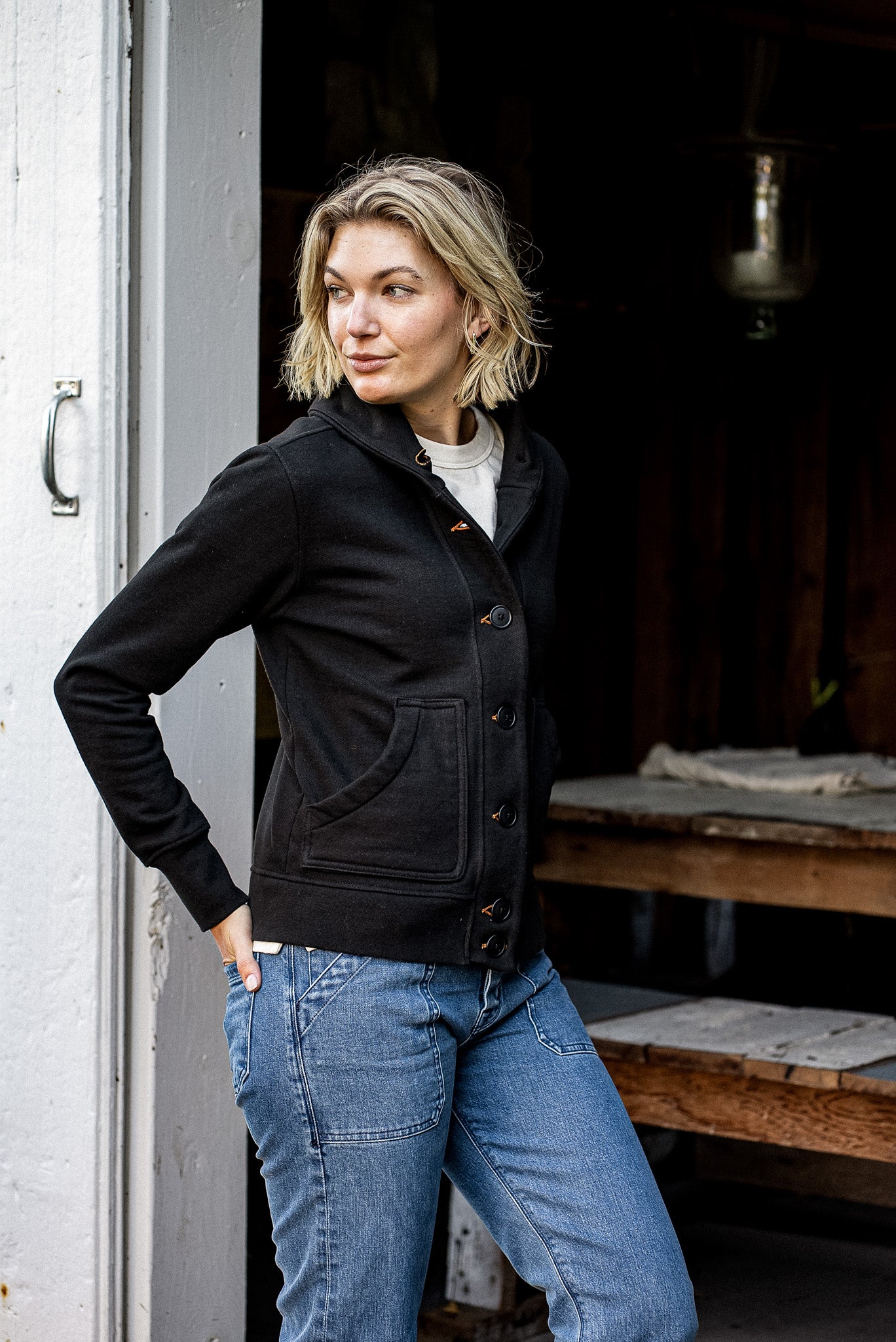Woman in a black French terry shawl cardigan with button front and patch pockets, paired with blue jeans.