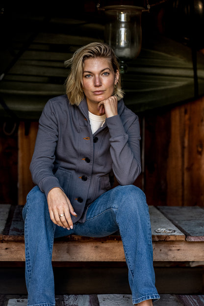 Woman in pebble grey French terry Bodega cardigan and blue jeans seated on a wooden porch.