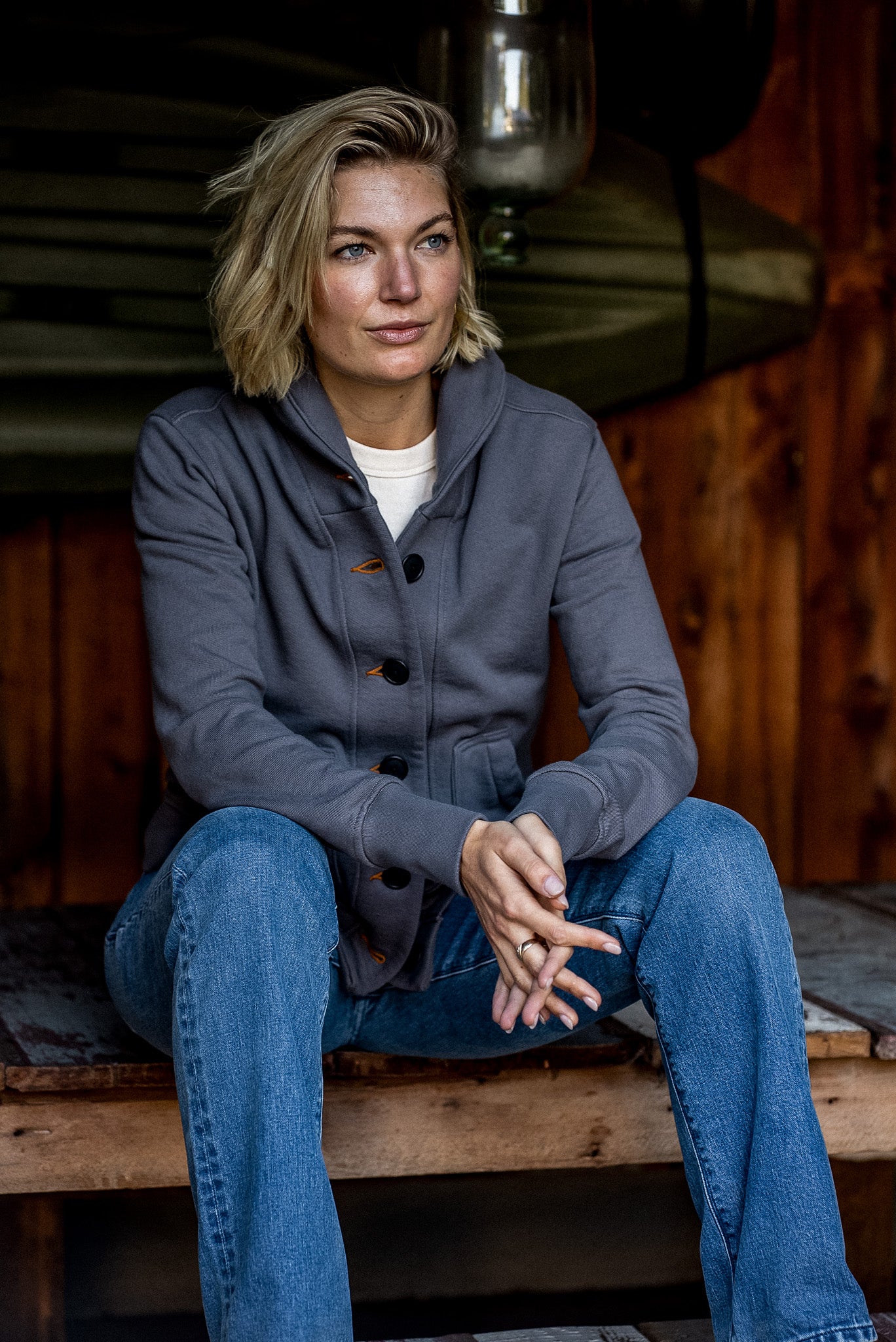 Woman seated on a wooden bench wearing the pebble gray Bodega shawl-collar French terry cardigan with tobacco keyhole.