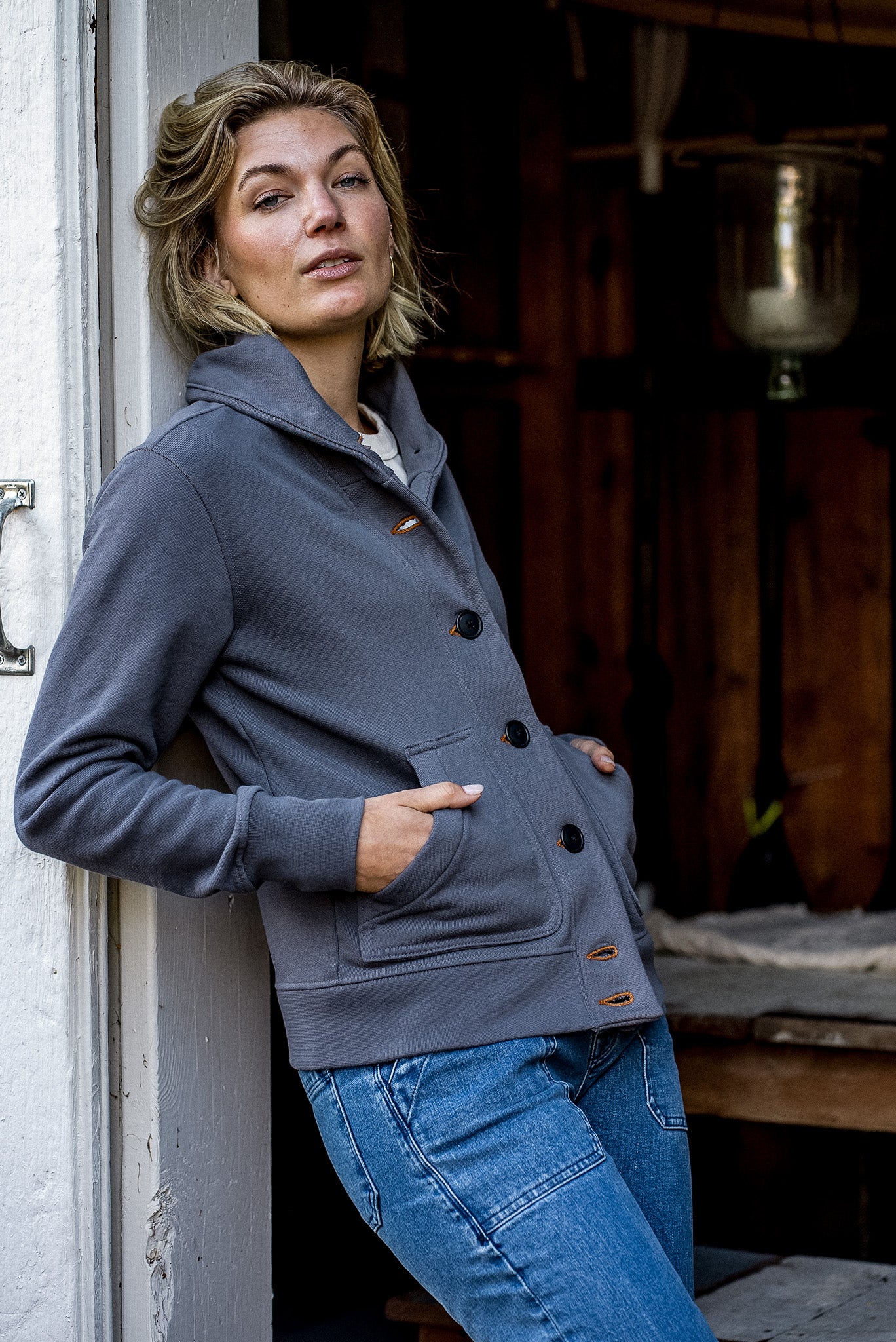 Person leaning in a doorway wearing the pebble grey Bodega shawl-collar French terry cardigan with tobacco keyhole.