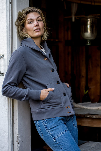 Person leaning in a doorway wearing the pebble grey Bodega shawl-collar French terry cardigan with tobacco keyhole.