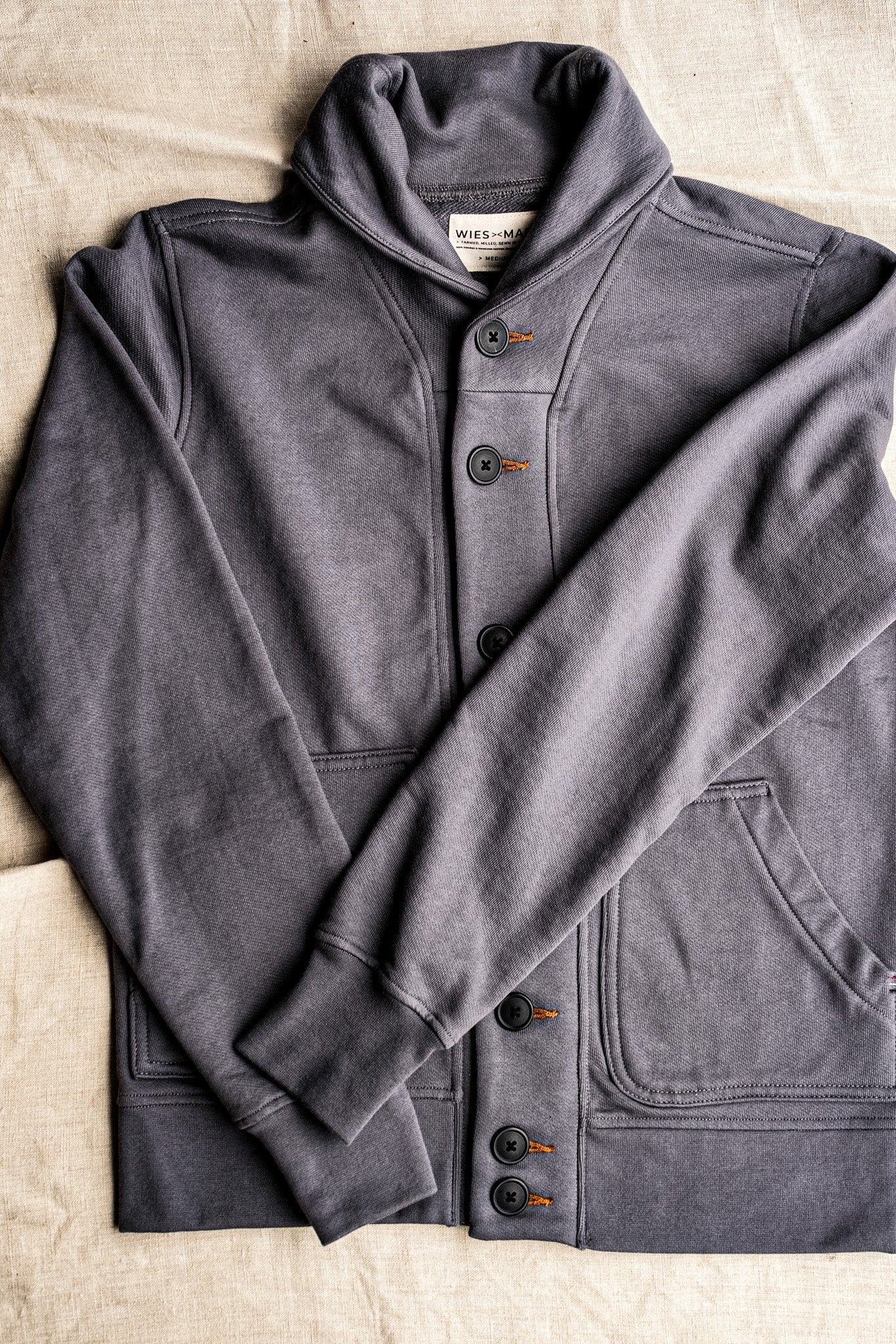 Pebble grey French terry shawl collar cardigan with black buttons and contrast tobacco keyhole stitching, laid flat on linen.
