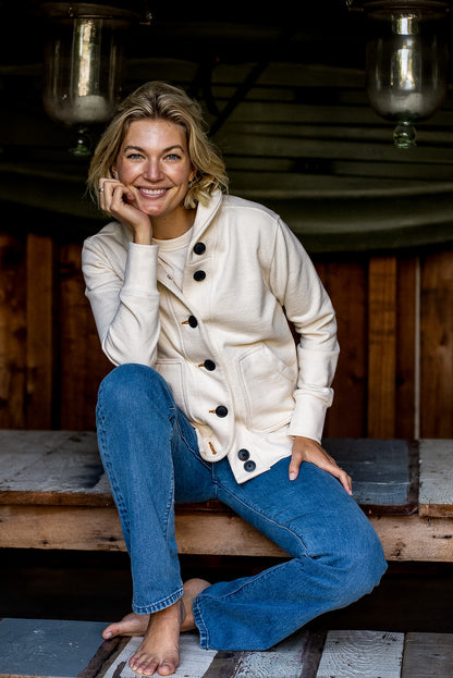 Woman in natural cotton French terry Bodega cardigan with contrast buttons, seated on wood bench.