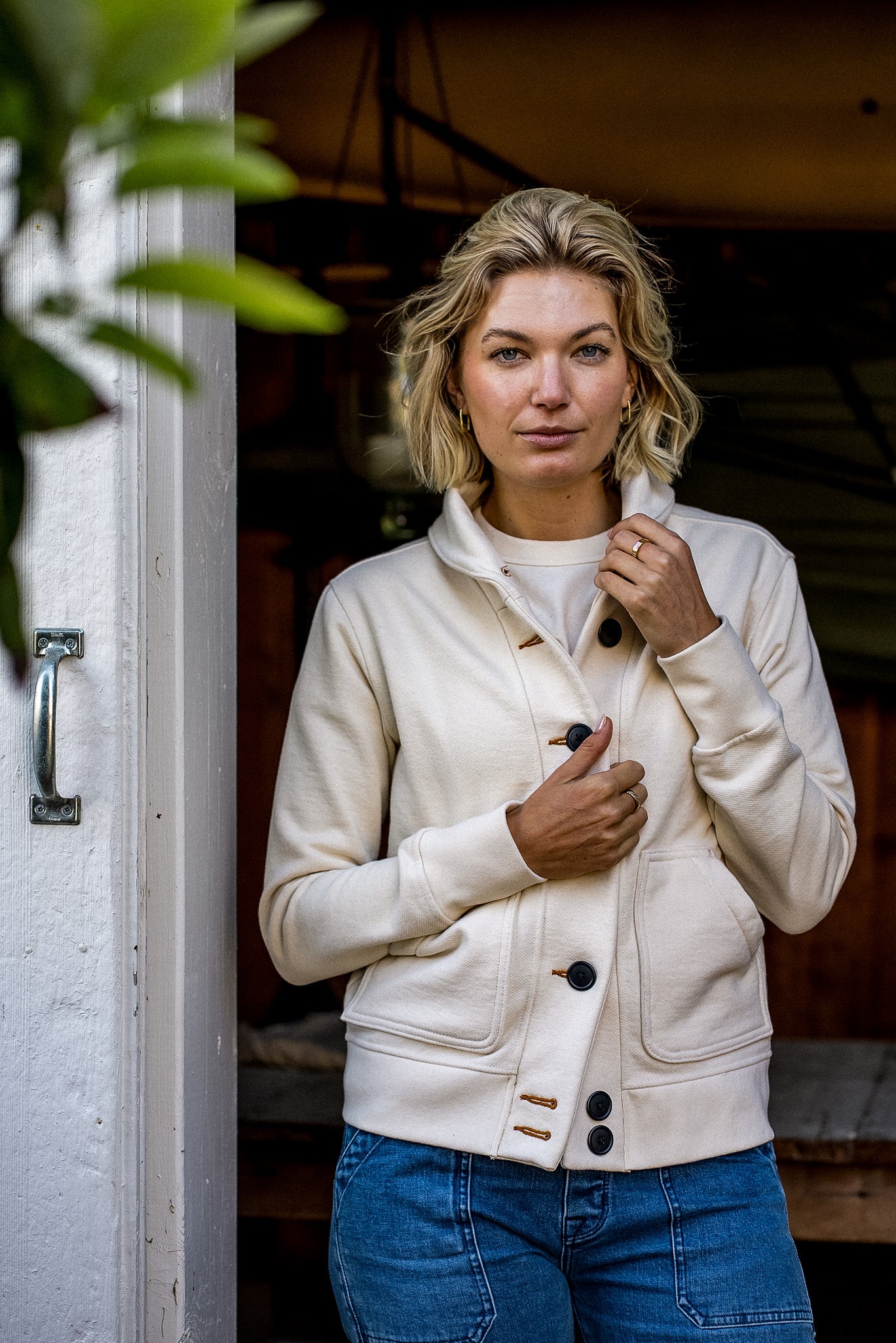 Model in a natural French terry cardigan with black buttons, standing in a wooden doorway.