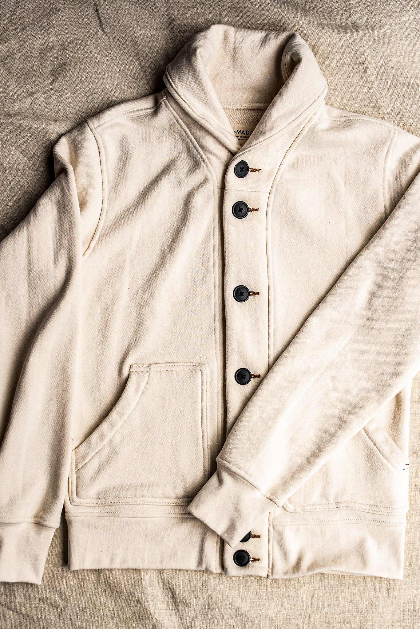 Natural French terry shawl-collar Bodega cardigan laid flat, showing dark buttons and front pocket.