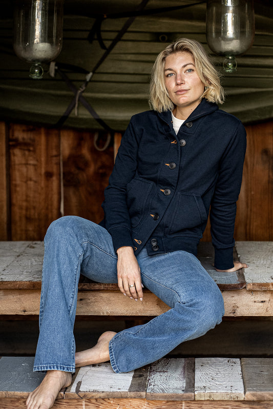Woman wearing navy Bodega cotton French terry cardigan with tobacco keyholes, seated on wood.