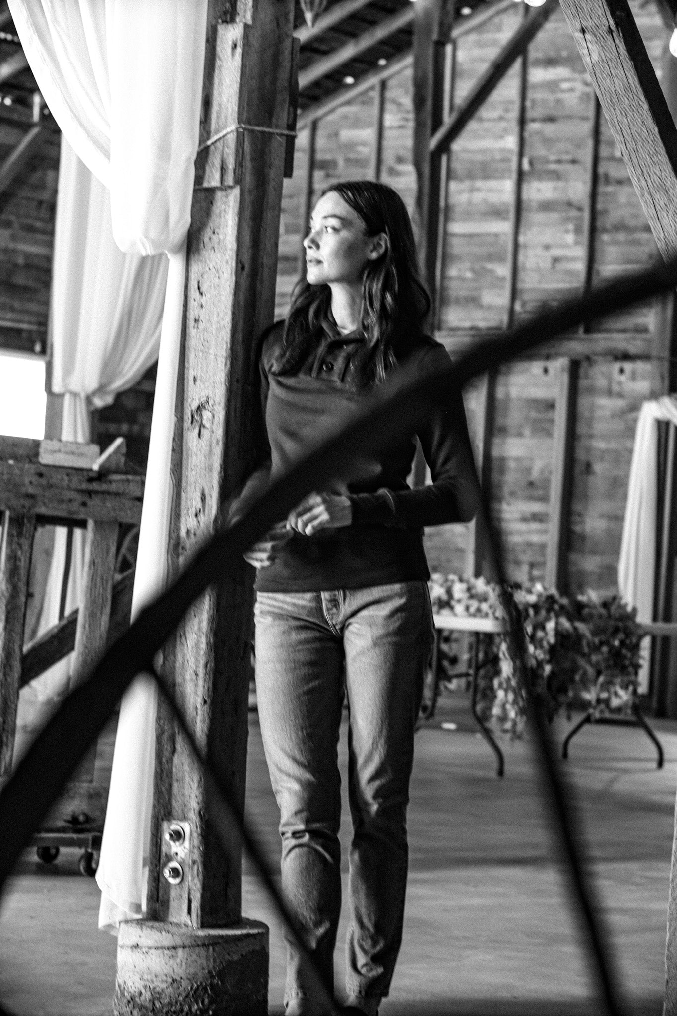 Woman in a Bodega shawl collar pullover and jeans standing in a rustic wooden mill, black and white.