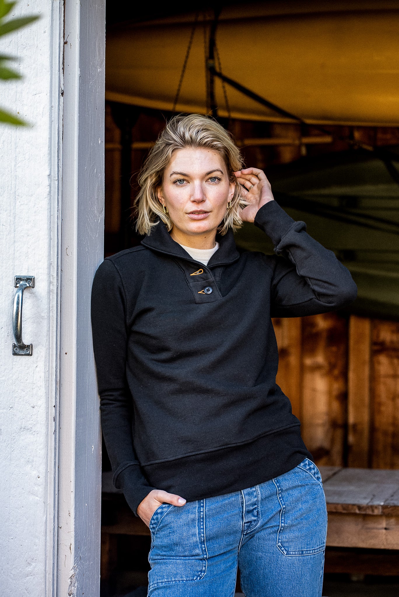 Woman in a black French terry shawl collar pullover and blue jeans leaning in a doorway.