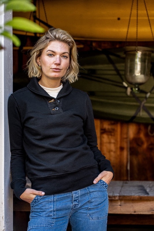 Women's Bodega shawl collar pullover in black French terry, worn with blue jeans on a wooden porch.