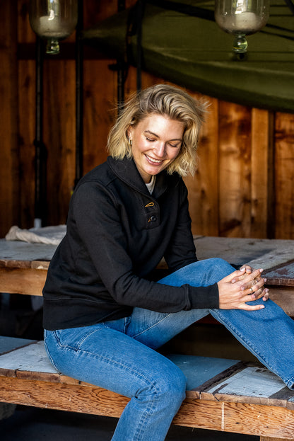 Woman in black Bodega shawl collar French terry pullover and blue jeans seated on a wooden bench.