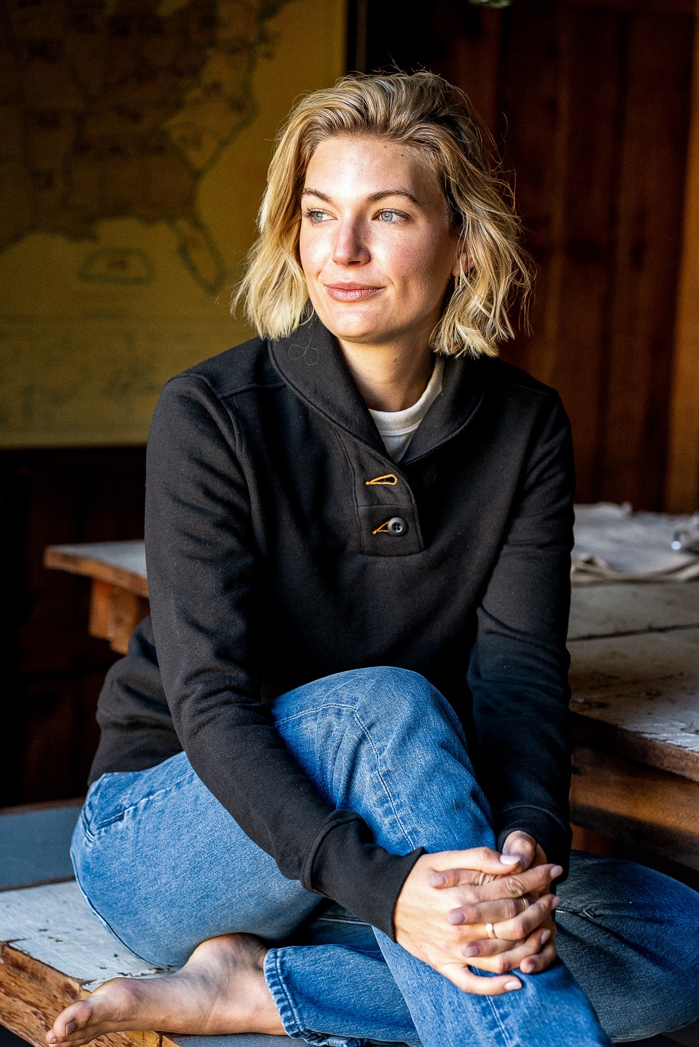 Woman in a black Bodega shawl collar pullover and blue jeans seated by a sunlit window.
