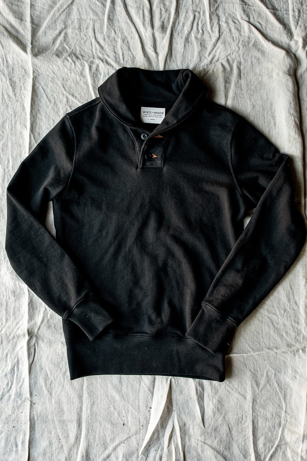 Black French terry shawl collar pullover laid flat on canvas, showing two-button placket and ribbed cuffs.