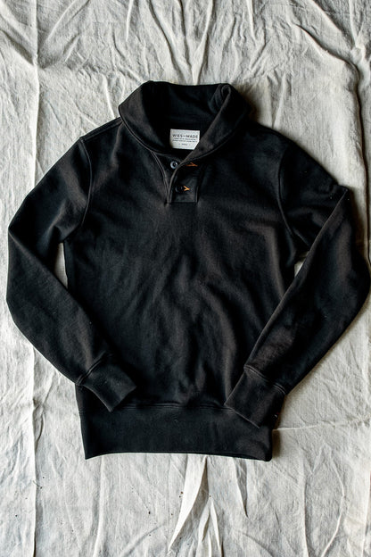 Black French terry shawl collar pullover laid flat on canvas, showing two-button placket and ribbed cuffs.