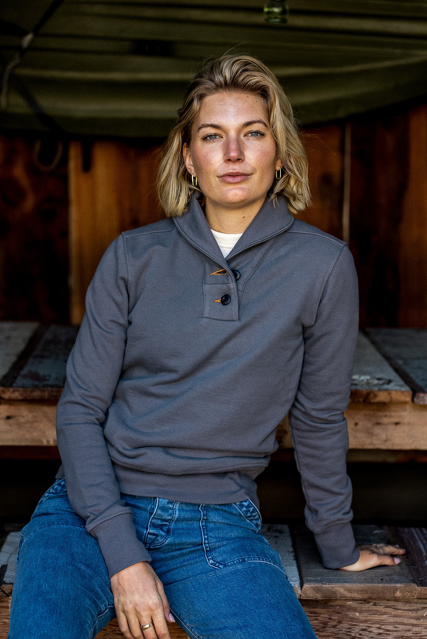 Woman wearing pebble gray Bodega shawl collar French terry pullover, seated on rustic wood.