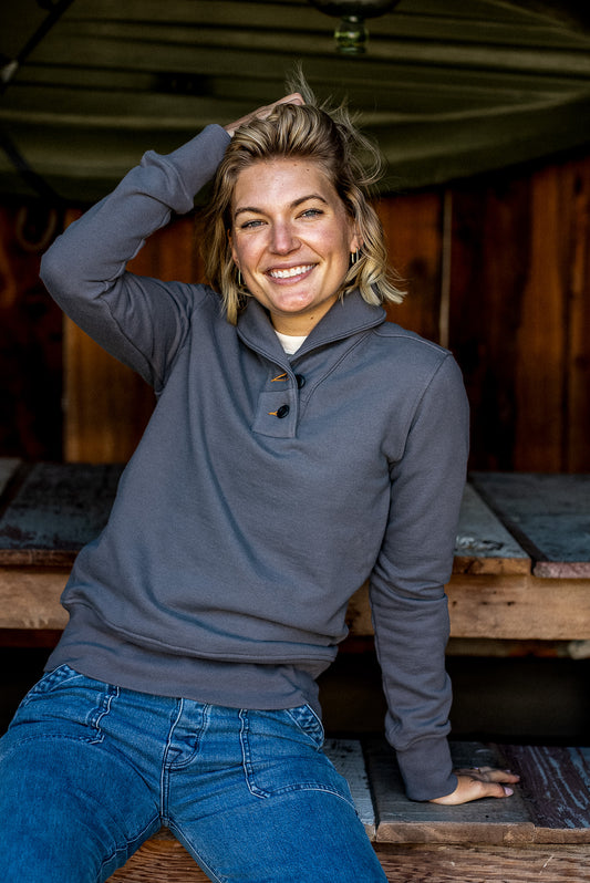 Smiling person wearing the Pebble gray Bodega French terry pullover, seated outdoors on wood.
