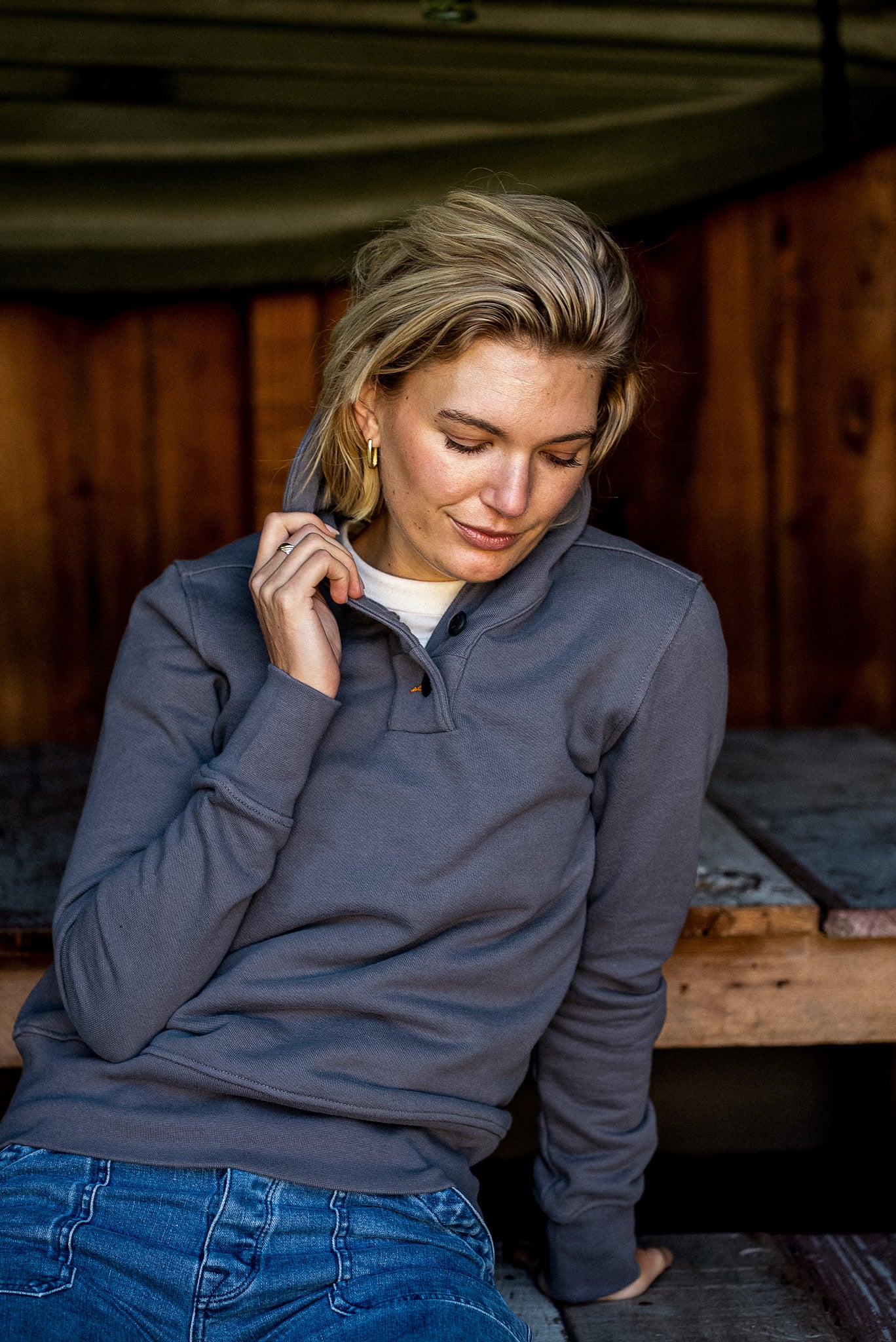 Person in pebble gray Bodega shawl pullover with snap placket, seated by wood cabin.