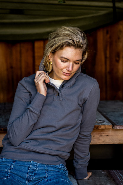 Person in pebble gray Bodega shawl pullover with snap placket, seated by wood cabin.