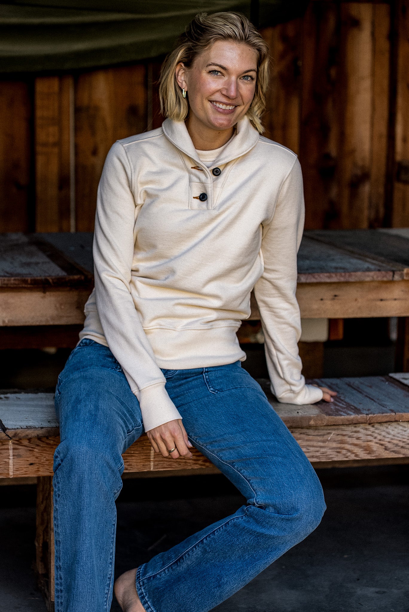 Woman in a natural cream French terry shawl-collar button pullover and blue jeans seated on a wooden bench.