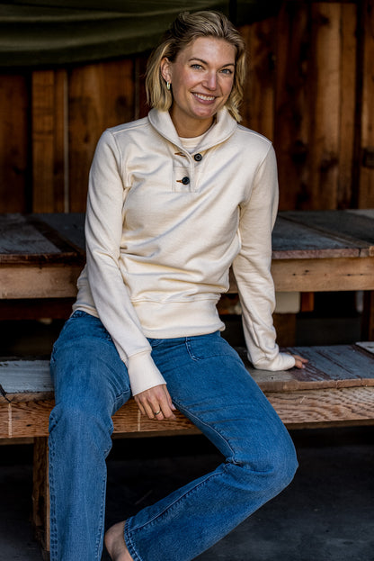 Woman in a natural cream French terry shawl-collar button pullover and blue jeans seated on a wooden bench.
