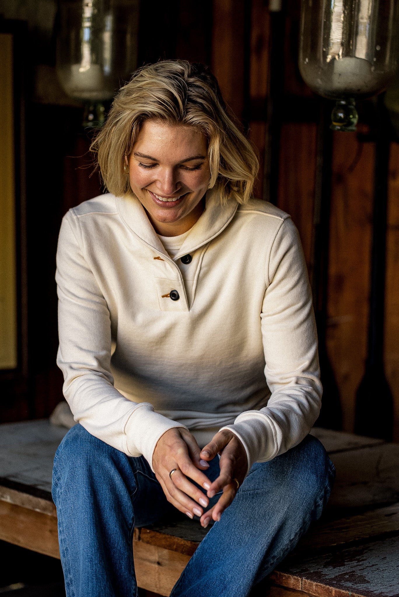 Woman in a natural cotton French terry shawl collar pullover and blue jeans, seated in a rustic wooden interior.