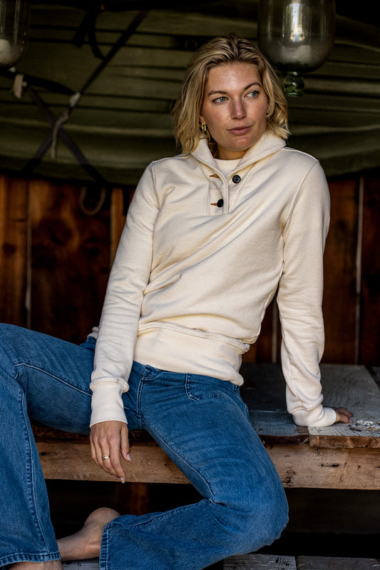 Woman in a natural cotton French terry shawl collar pullover and blue denim jeans, seated on a wooden bench.