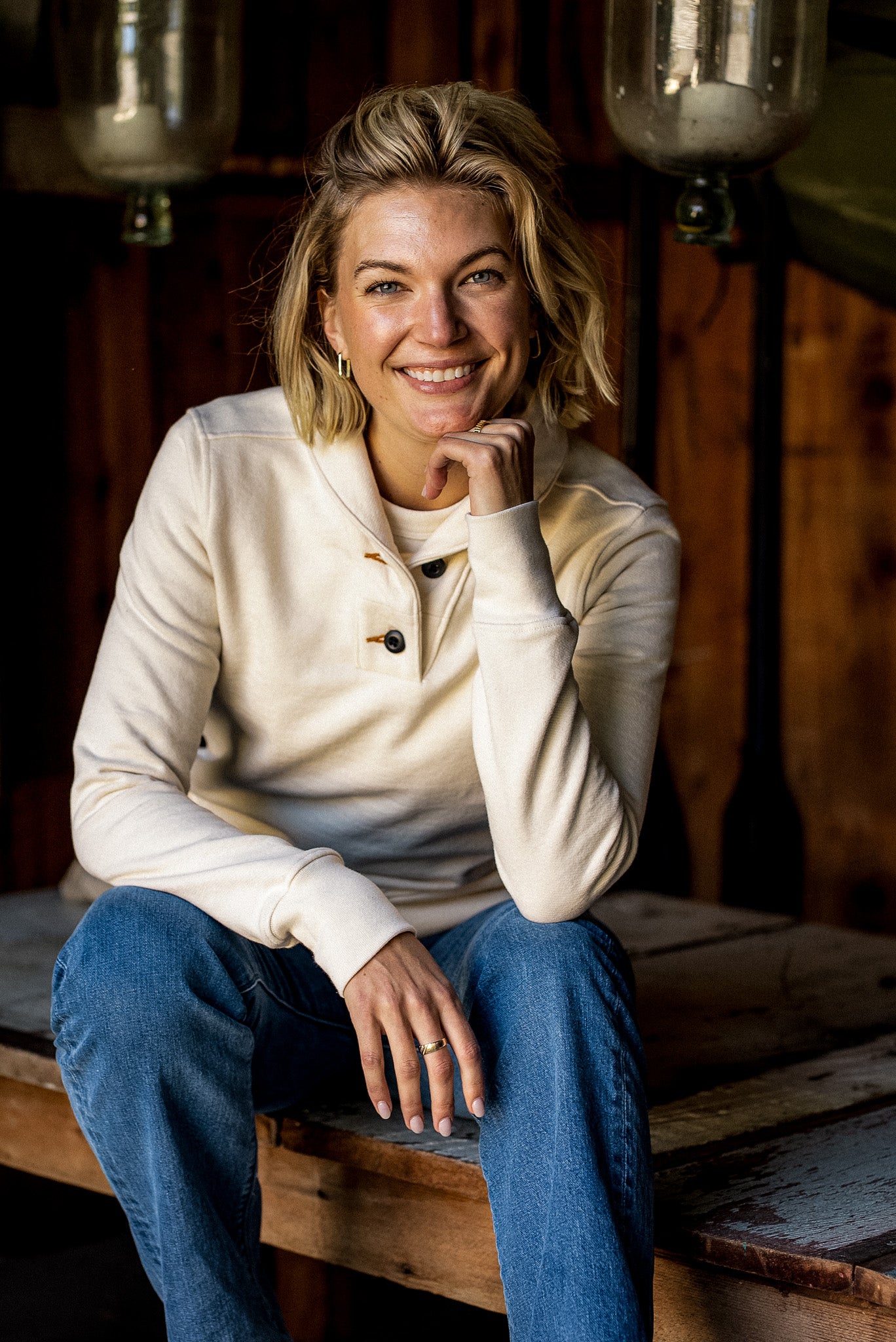 Smiling model in a natural cream French terry shawl pullover and blue jeans, seated in a rustic wood cabin.