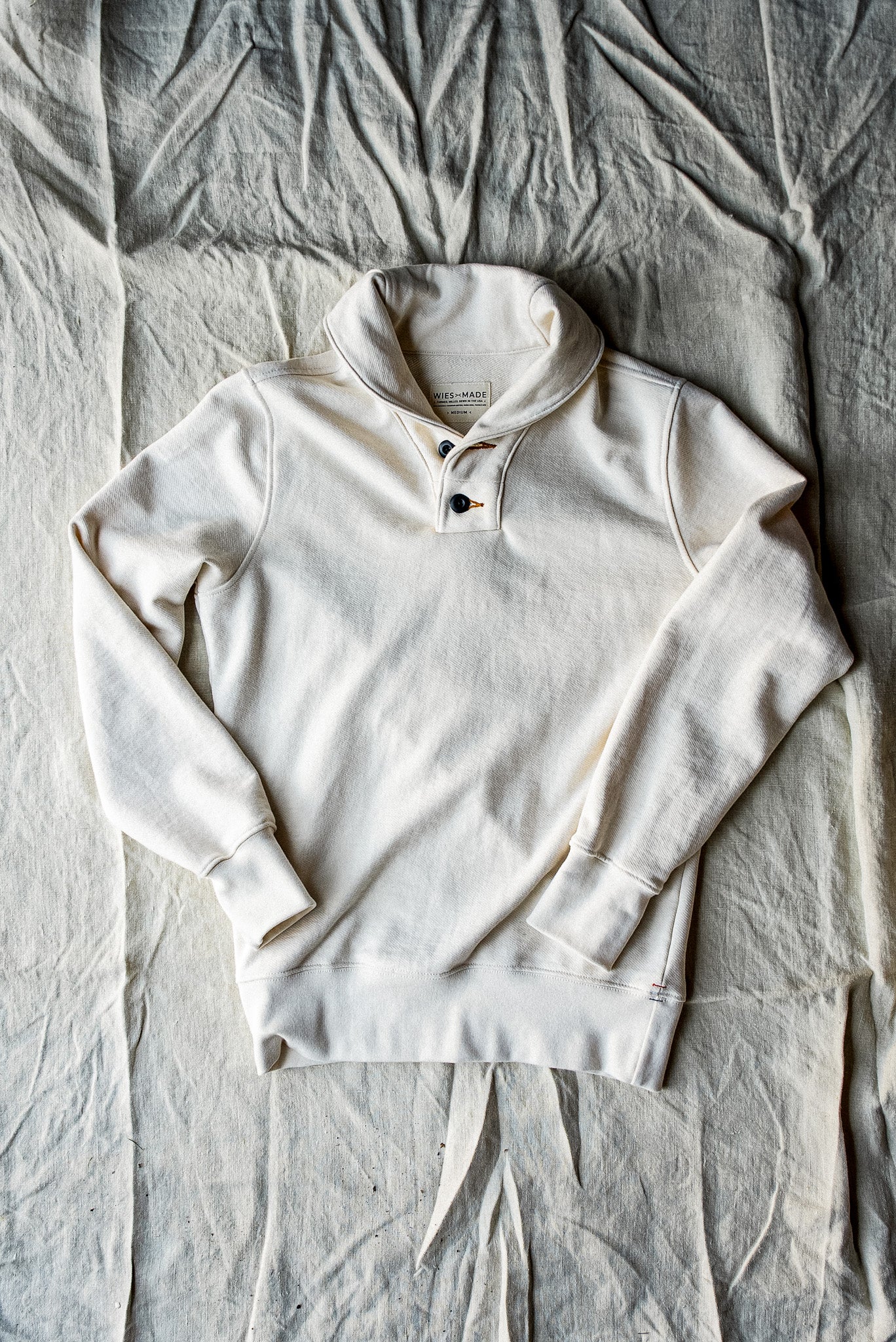 Natural cotton French terry shawl collar pullover with two-button placket, laid flat on canvas.