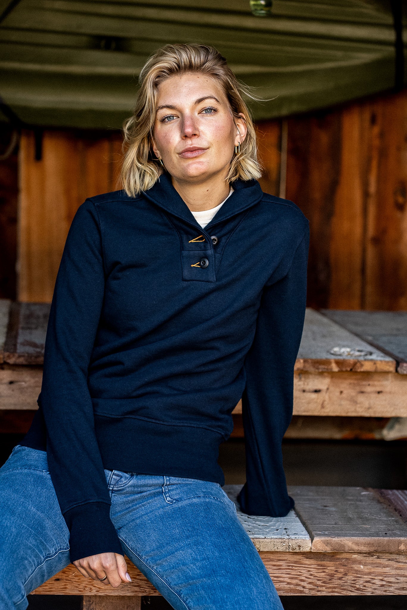 Woman wearing navy Bodega shawl collar French terry pullover with jeans, seated on rustic wood.