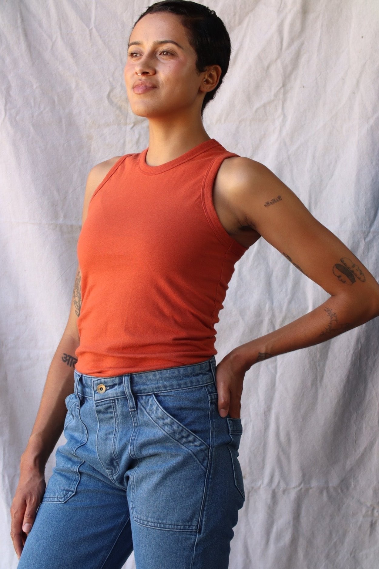 Model wearing a brick red Bolinas cotton tank tucked into blue denim jeans against a linen backdrop.