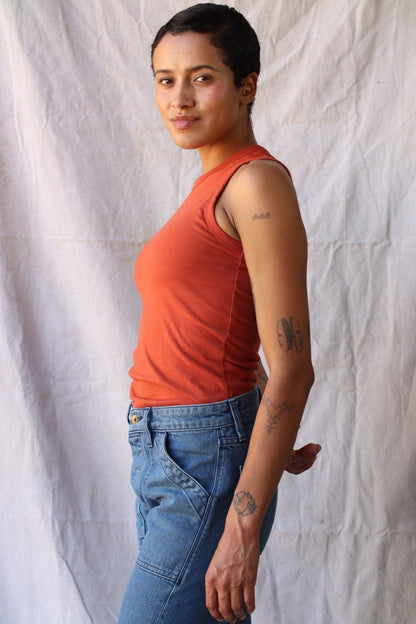 Side view of the brick-colored Bolinas cotton tank tucked into blue jeans against a canvas backdrop.
