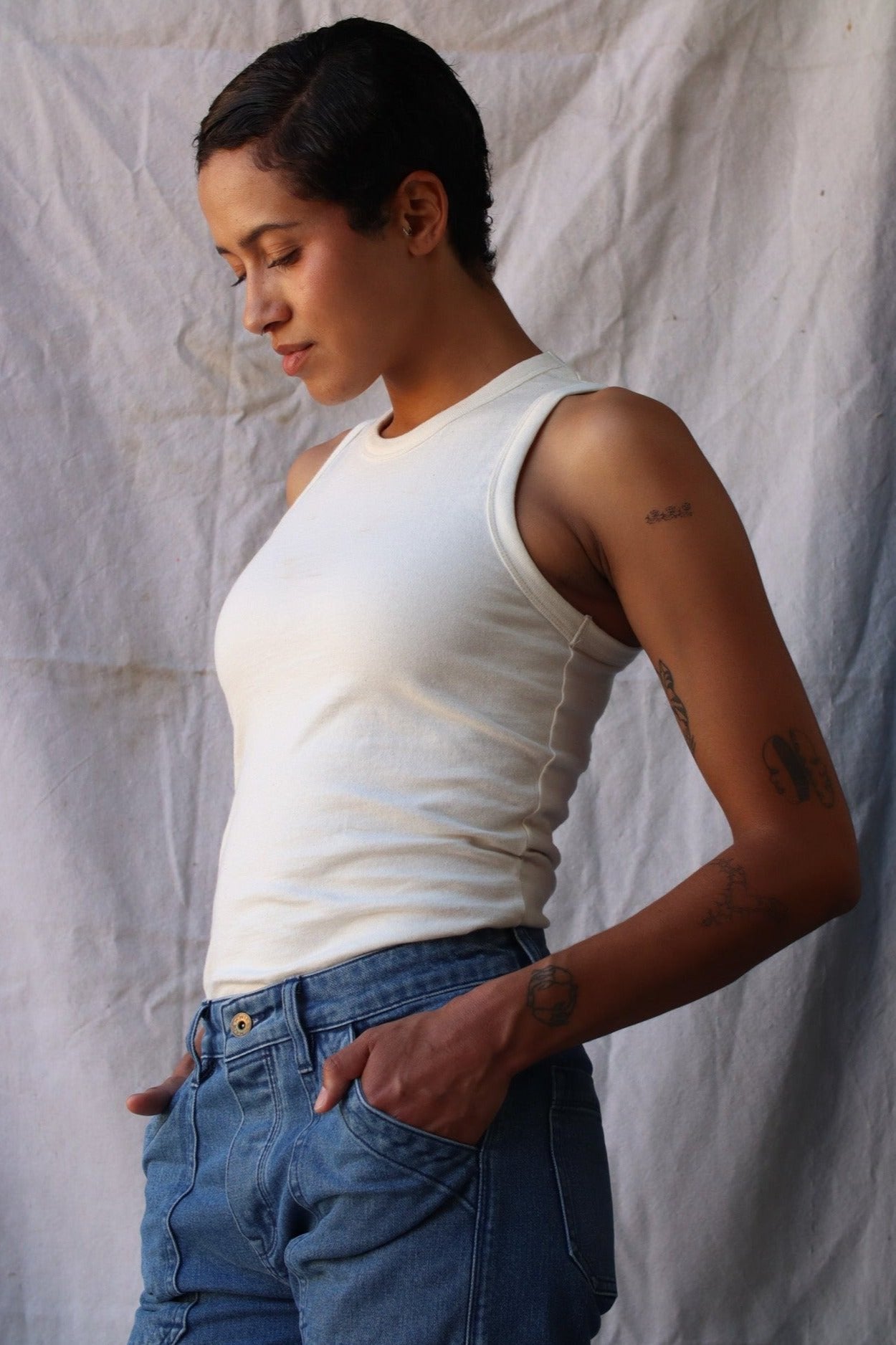Model wears the natural cotton Bolinas tank tucked into blue jeans against a draped canvas backdrop.