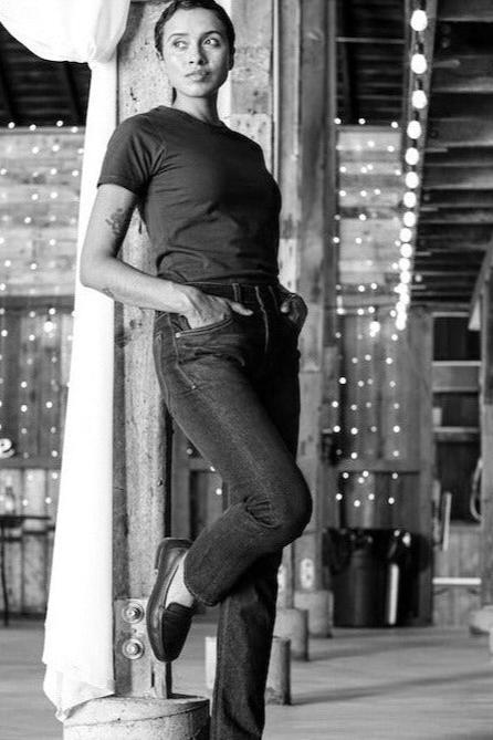 Model leaning against a post in the olive Bolinas heavyweight tee and dark jeans inside a string-lit barn.