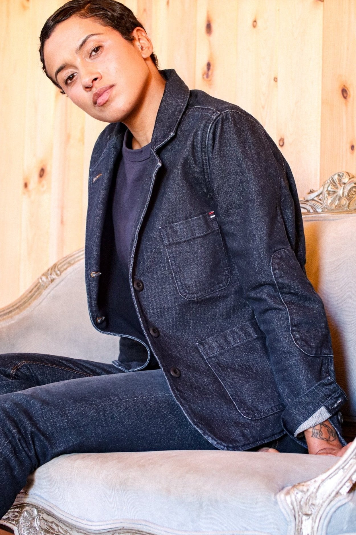 Model in a dark denim chore jacket and matching jeans seated on an ornate settee.
