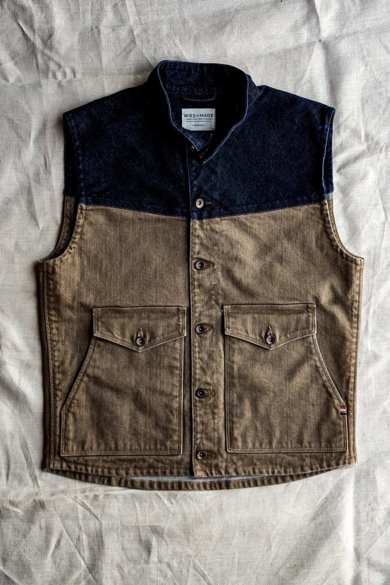 Broken twill denim vest with wheat-colored body, indigo yoke, and double button pockets laid on linen.