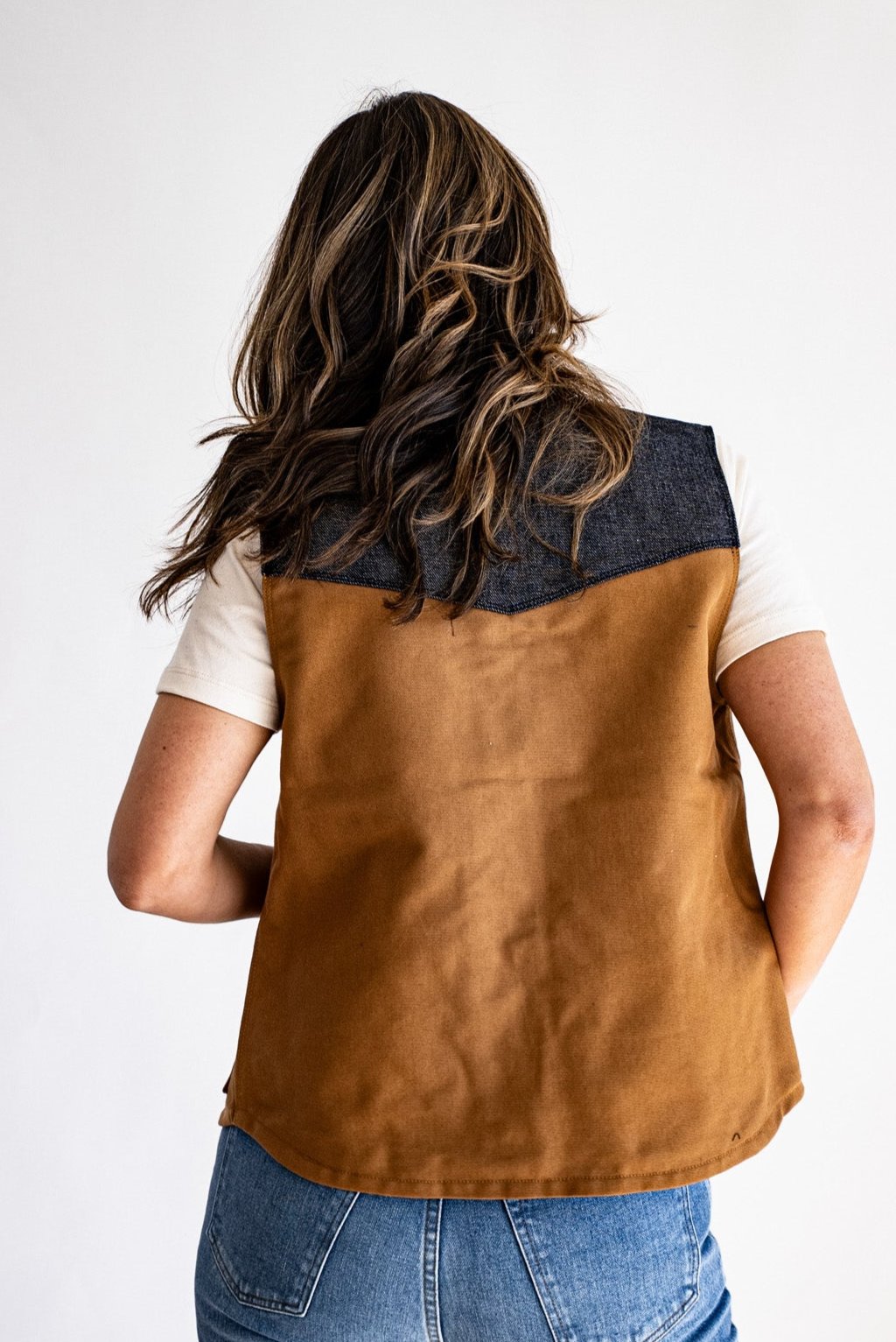 Back view of the Jamie vest in tan canvas with a raw denim yoke, worn over a cream tee and jeans.