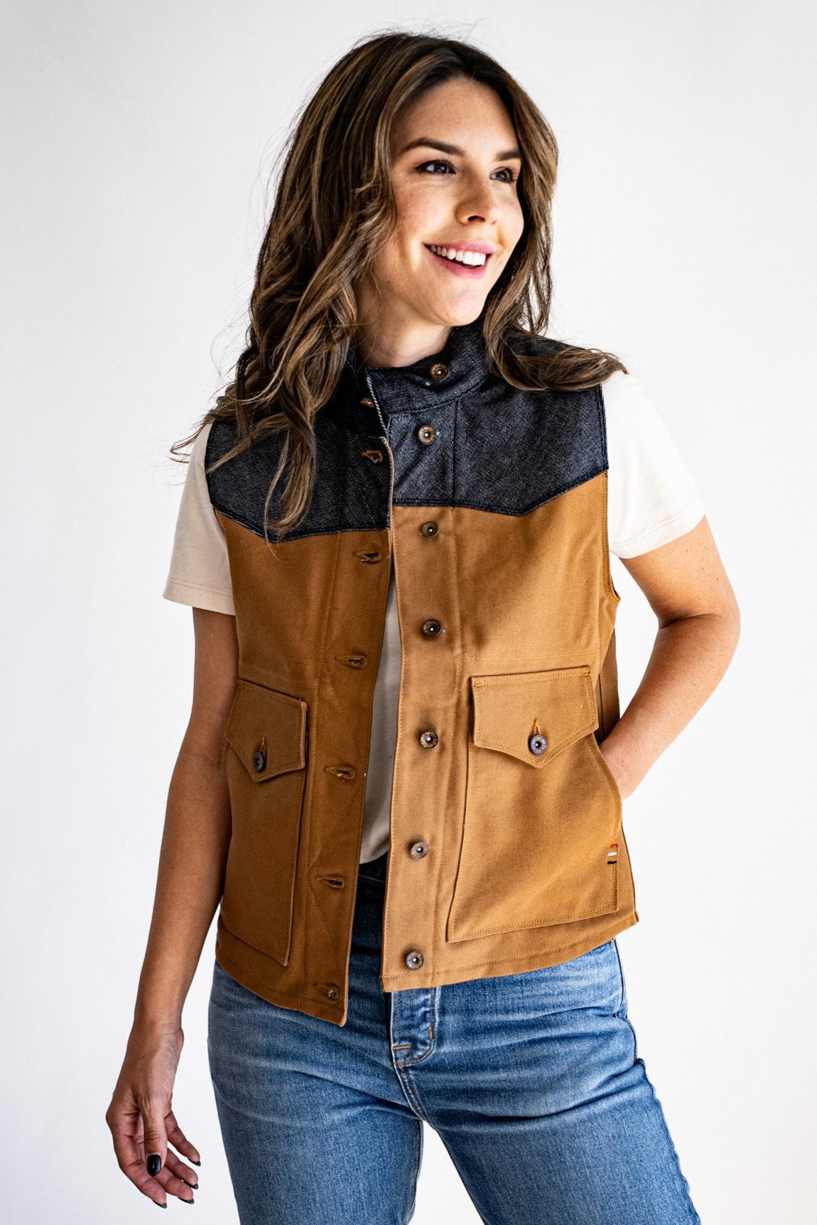 Woman wearing a tan canvas Jamie vest with denim yoke and brass buttons over a cream tee and jeans.