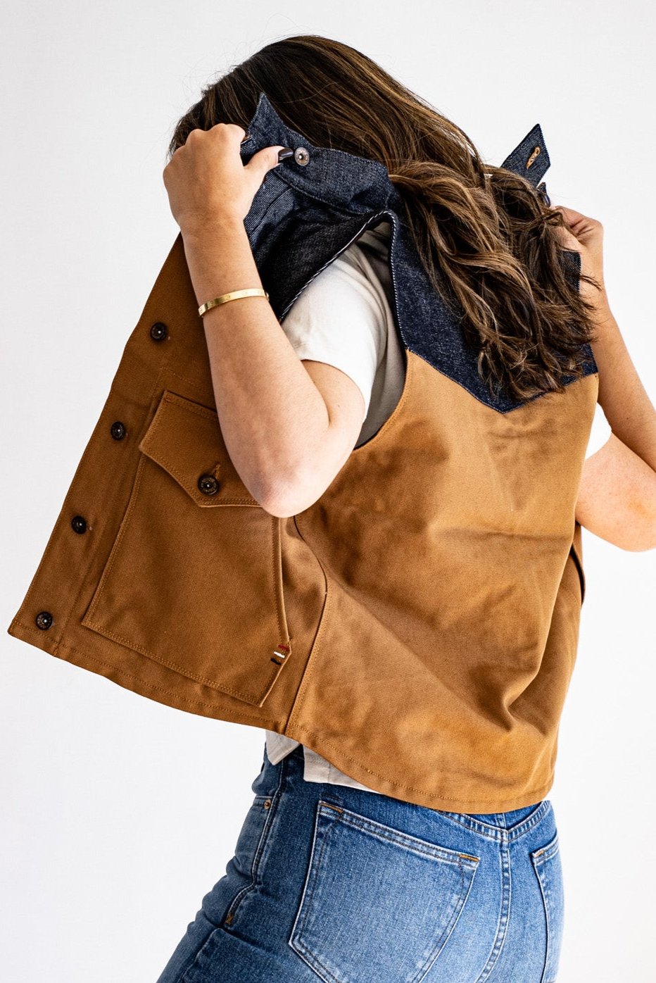Woman pulling on a tan canvas Jamie vest with denim-lined collar, button pocket, and brass hardware.