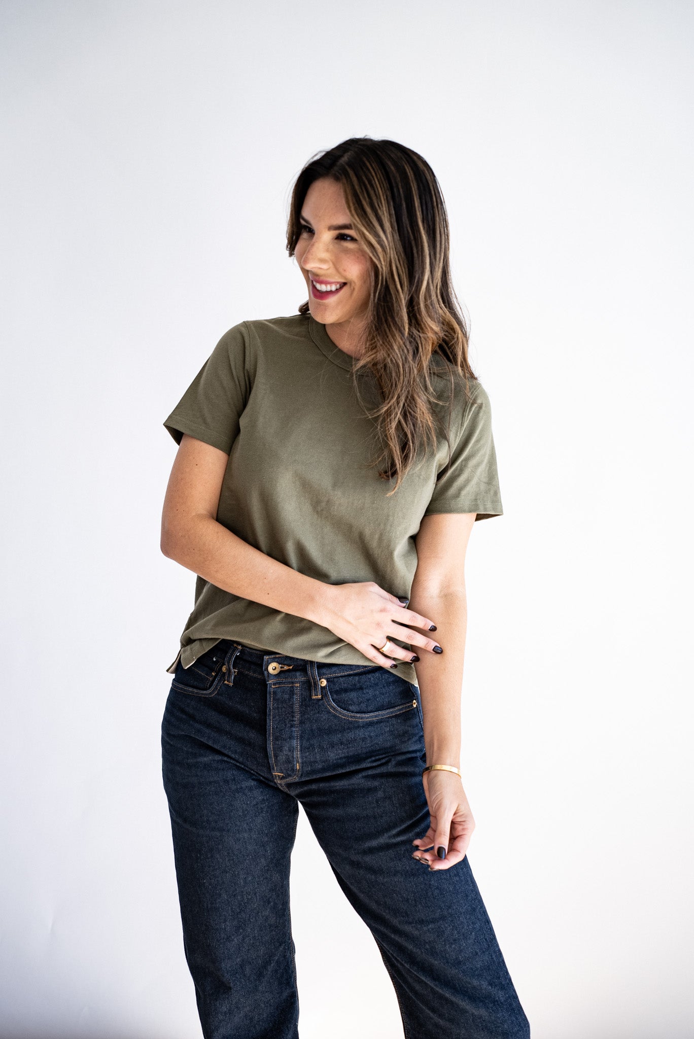 Woman wearing an olive boxy tee with dark raw denim jeans against a white studio backdrop.