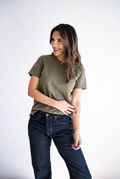 Woman wearing an olive boxy tee with dark raw denim jeans against a white studio backdrop.