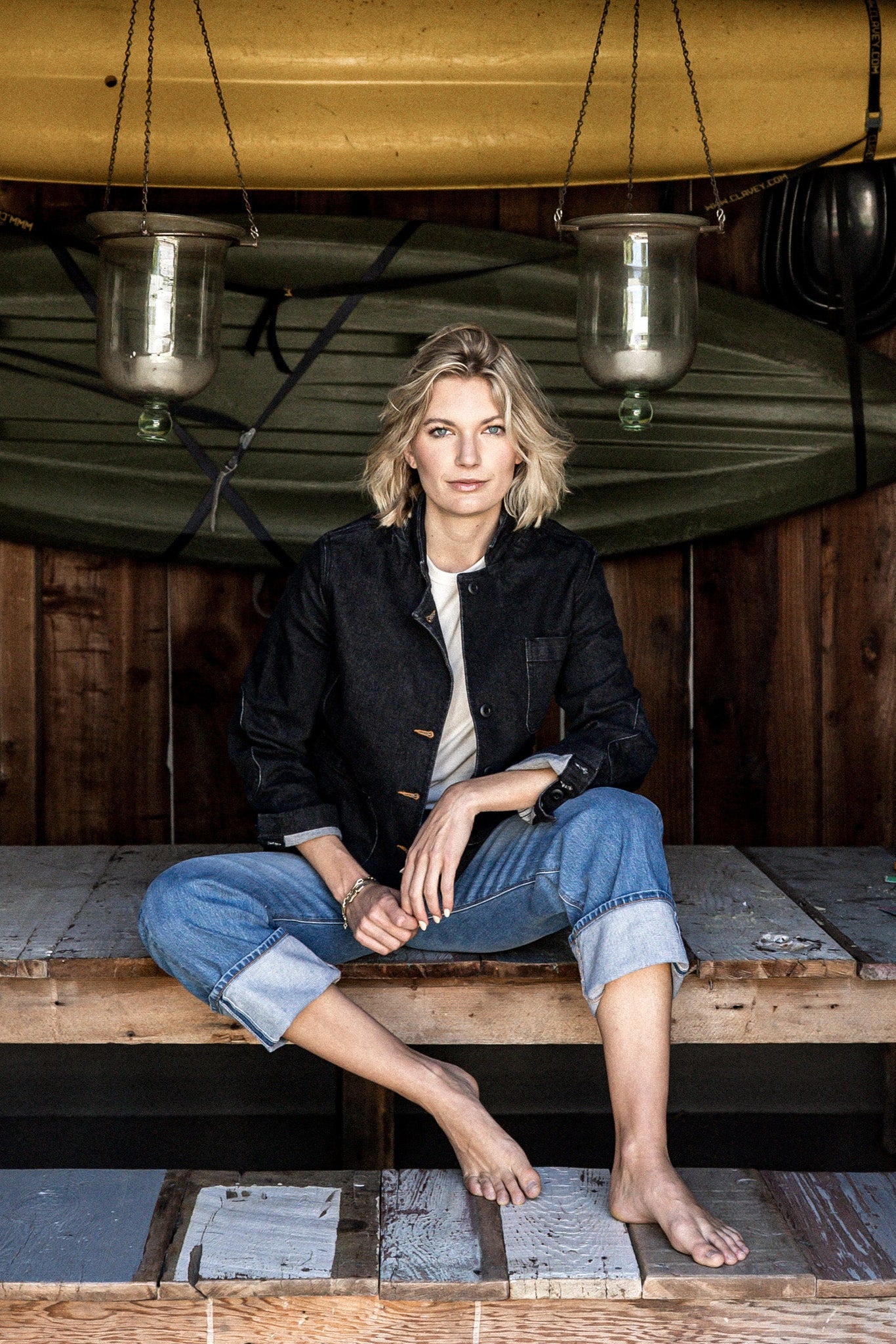 Woman in a black denim chore blazer and cuffed jeans seated on a wooden deck beneath a canoe.