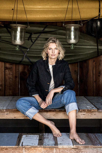 Woman in a black denim chore blazer and cuffed jeans seated on a wooden deck beneath a canoe.