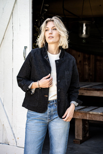 Woman wearing black denim chore blazer over a white tee with light-wash jeans, standing in a barn doorway.