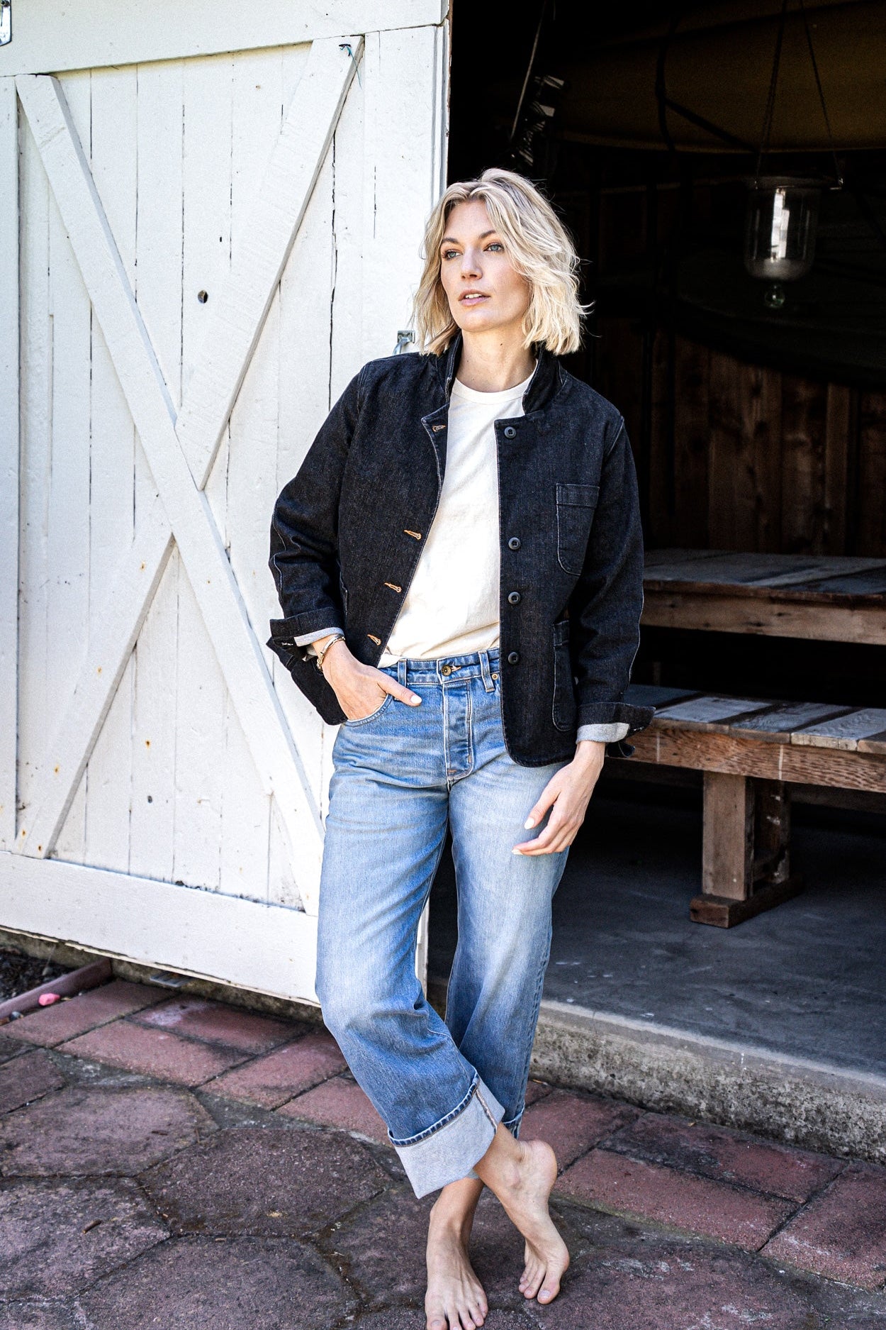 Woman in a black denim chore blazer, white tee, and cuffed light-wash jeans leaning against a white barn door.