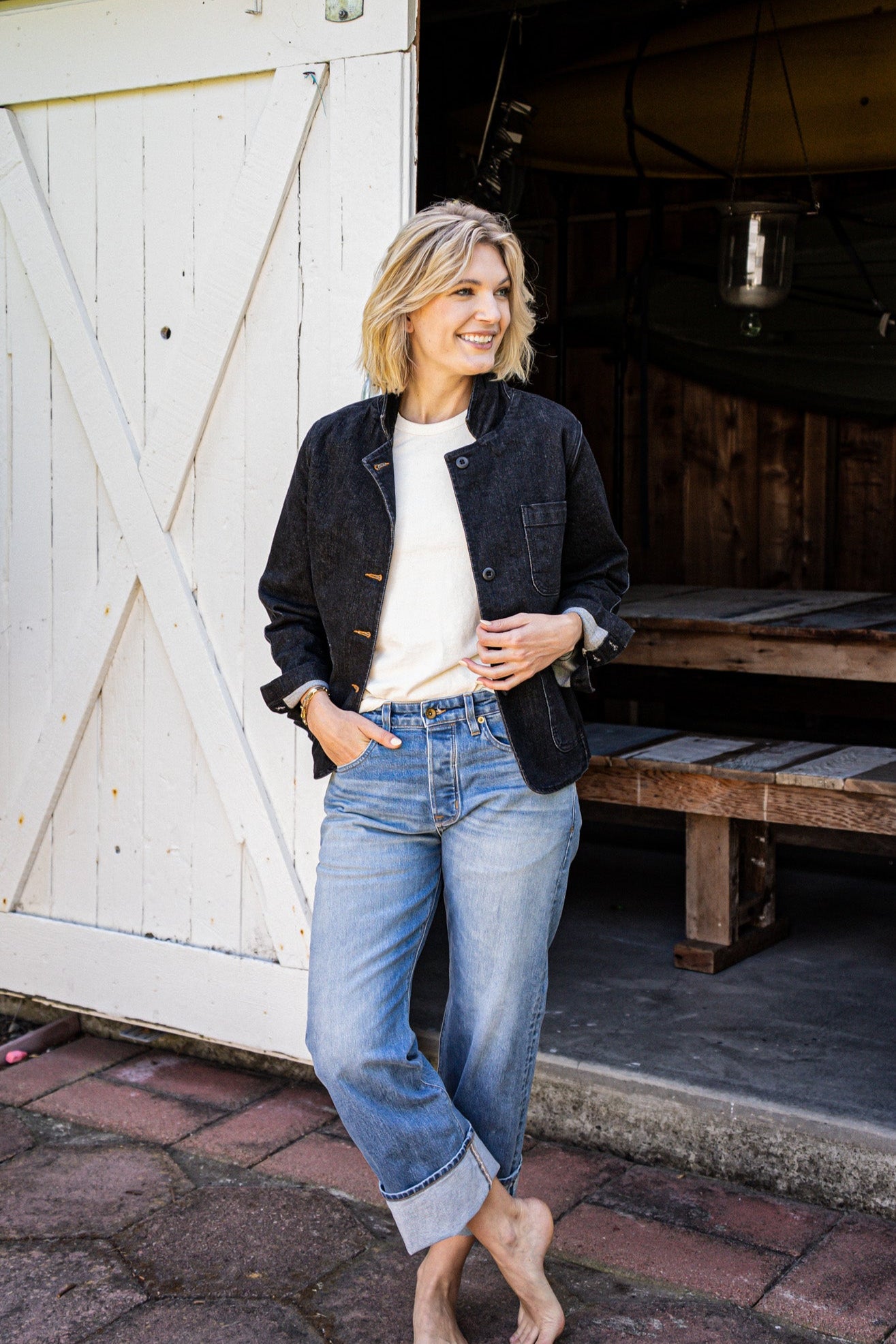 Woman in a black denim chore blazer over a cream tee and cuffed blue jeans, standing by a white barn door.
