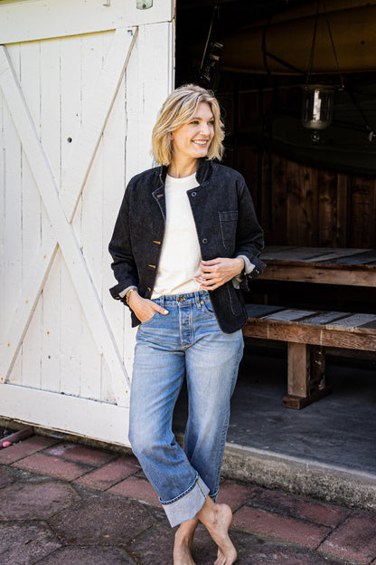 Woman in a black denim chore blazer over a cream tee and cuffed blue jeans, standing by a white barn door.
