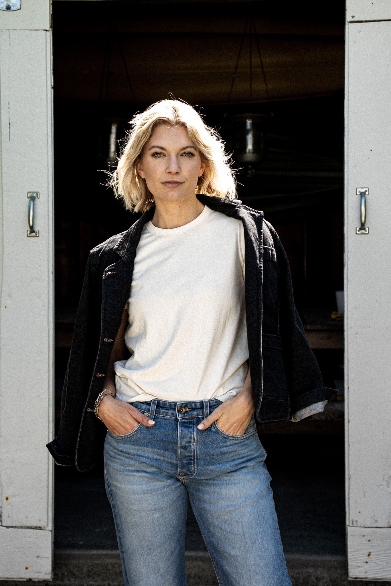 Woman wearing the black denim Scout Blazer draped over a cream tee with light wash jeans in a barn doorway.