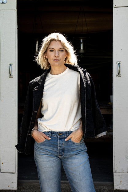 Woman wearing the black denim Scout Blazer draped over a cream tee with light wash jeans in a barn doorway.