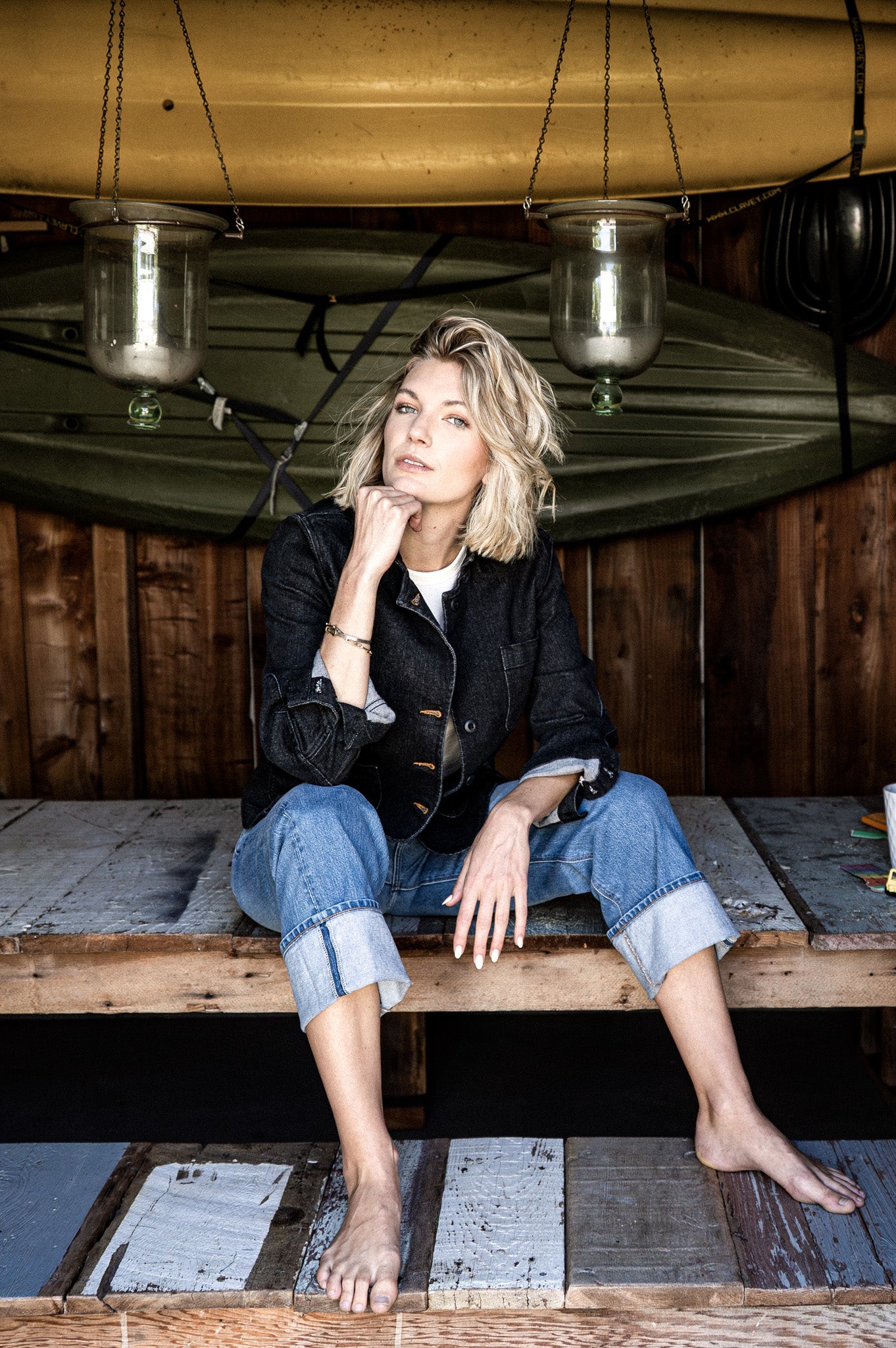 Woman wearing the black denim Scout chore blazer with cuffed jeans, seated on a wood platform beneath a hanging canoe.