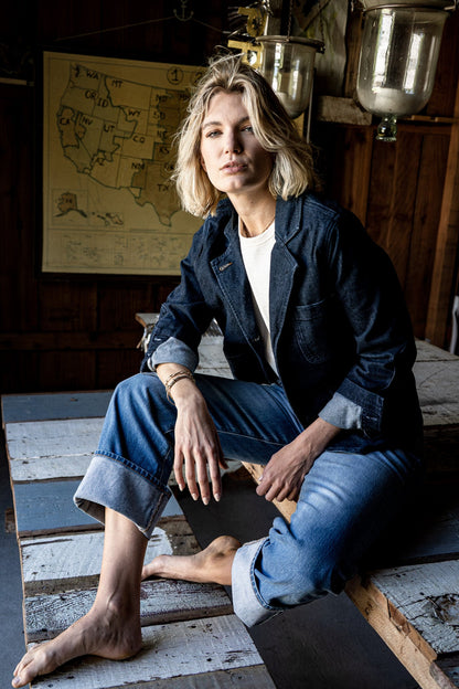 Woman in a dark indigo denim chore blazer and cuffed jeans seated on a wooden pallet near a US map.